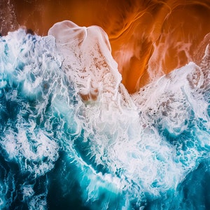 May include: An aerial view of ocean waves breaking on a sandy beach. The water is a deep blue with white foam, and the sand is a warm brown.