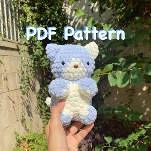 May include: A crocheted plush toy in the shape of a cat. The cat is white and light blue with a white face and blue ears. The cat is being held in a hand. The text "PDF Pattern" is written in blue letters on a green background.