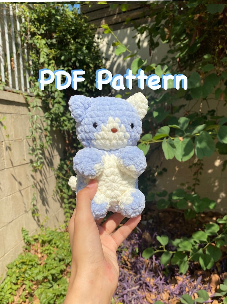 May include: A crocheted plush toy in the shape of a cat. The cat is white and light blue with a white face and blue ears. The cat is being held in a hand. The text "PDF Pattern" is written in blue letters on a green background.
