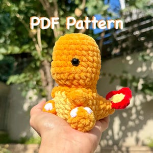 May include: A handmade, orange crochet creature with a black eye and a red and yellow tail. The text "PDF Pattern" is displayed above the creature. The creature is held in a hand.