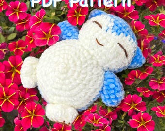 May include: A handmade, white and blue crochet plushie in the shape of a sleeping animal, resting among vibrant pink flowers. The text "PDF Pattern" is displayed above the plushie. The plushie has blue accents and closed eyes.