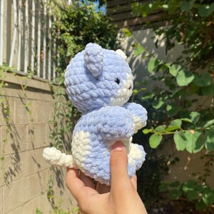 May include: A crocheted plush toy in the shape of a bear. The bear is light blue and white with a fluffy texture. The bear is being held in a hand.