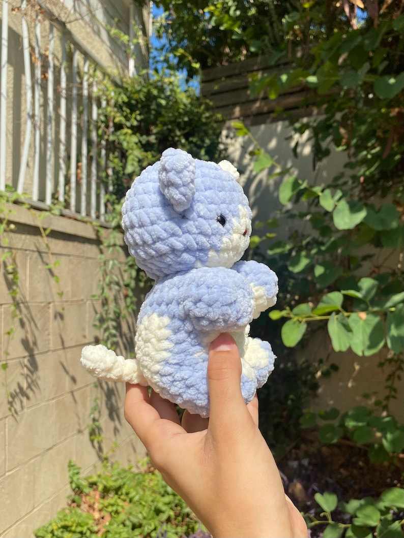 May include: A crocheted plush toy in the shape of a bear. The bear is light blue and white with a fluffy texture. The bear is being held in a hand.