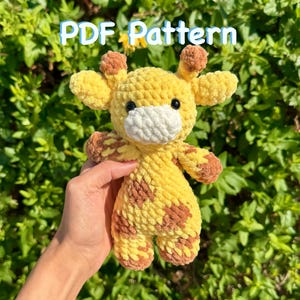 May include: A handmade, plush giraffe toy in yellow and brown. The giraffe has a white muzzle, brown spots, and small brown horns. The text "PDF Pattern" is displayed in a light blue font above the giraffe.