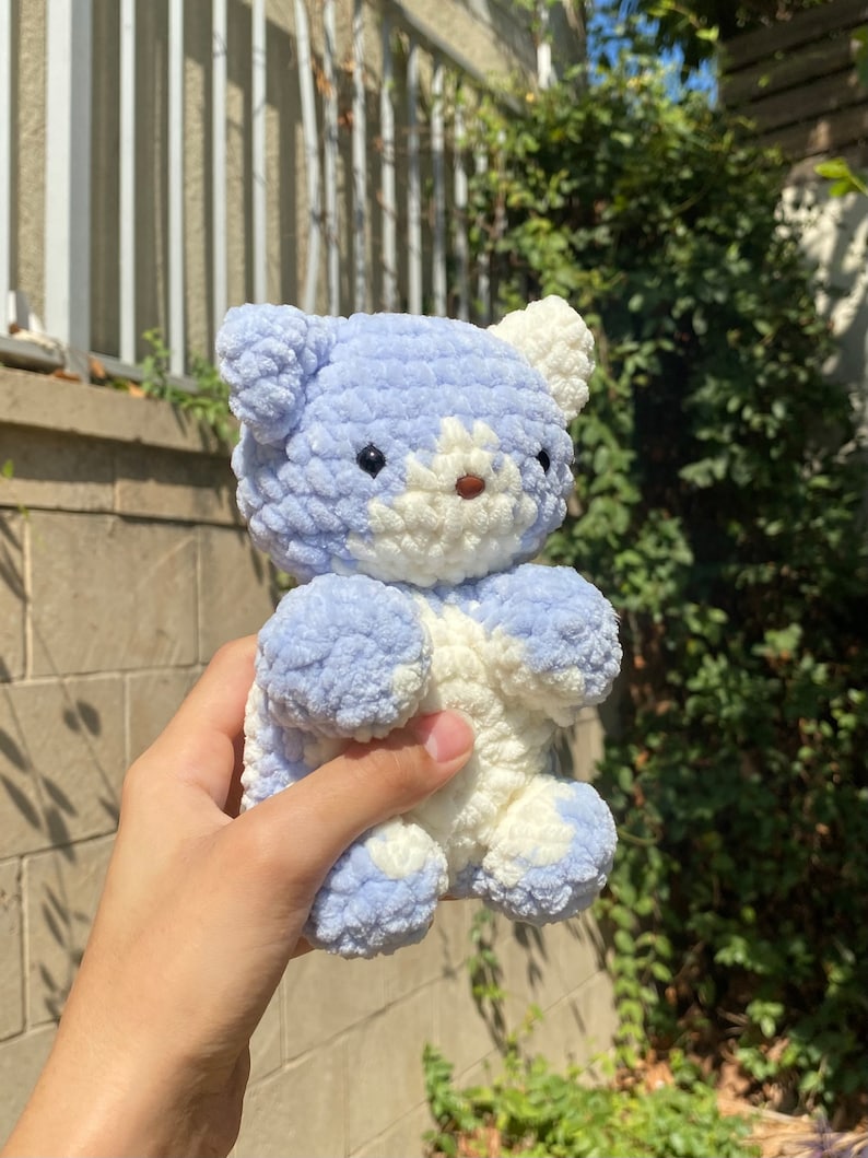 May include: A small, crocheted plush toy bear in a light blue and white colour scheme. The bear has a white face with black eyes and a pink nose.
