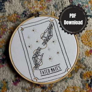 May include: An embroidery hoop featuring a white fabric with a black and gold design. The design includes two skeleton hands connected by a gold chain, stars, and the words "FATED MATES". A black circle with "PDF Download" is in the upper right.