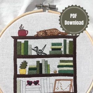 May include: Embroidered design of a bookshelf with a cat sleeping on top, a mug of coffee, a cactus, books, a origami crane, sunglasses, and a framed picture. The design is stitched on a white fabric.