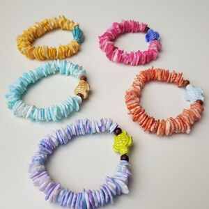 Puede incluir: Cinco pulseras coloridas hechas de pequeñas piezas planas con forma de concha. Cada pulsera tiene un pequeño dije de tortuga de un color diferente.