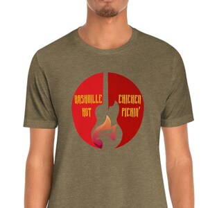 以下が含まれることがあります： 赤い円形のグラフィックが描かれた緑色のTシャツ。グラフィックは、ギターのシルエットで、左側に「Nashville Hot」、右側に「Chicken Picin'」というテキストが書かれています。