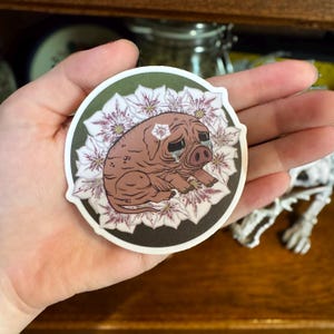 May include: A round sticker featuring a brown pig with a sad expression, tears, and a flower on its head. The pig is surrounded by white and purple flowers, set against a dark green background.
