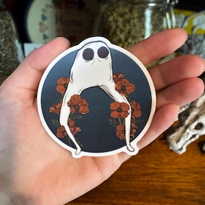 May include: A round sticker featuring a white ghost-like figure with two black eyes, surrounded by orange poppy flowers against a dark blue background. The sticker is held in a hand.