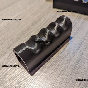 Op de afbeelding: Een zwart 3D-geprint object met een golvend, getextureerd oppervlak. Het object heeft de vorm van een rechthoekig prisma met een gebogen bovenkant.