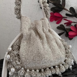 Puede incluir: Un bolso plateado con cuentas y cordón, con asa de terciopelo blanco. El bolso está adornado con perlas y flecos de cuentas. Un bolso pequeño y elegante, un accesorio con estilo.