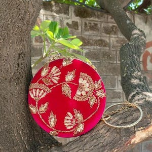 Peut inclure: Pochette ronde rouge avec broderies florales dorées. La pochette est ornée d'une chaîne dorée et est présentée sur une branche d'arbre. Le mot "Red" est dans un rectangle blanc en arrière-plan.