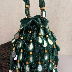 Pode incluir: Uma bolsa de cordão de veludo verde escuro com detalhes decorativos de espelho em forma de gota e ferragens douradas. A bolsa tem uma textura de contas e alças verdes combinando. A bolsa está em uma cesta de vime.