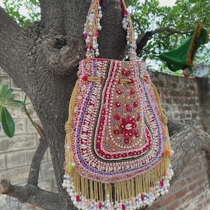 May include: A pink and red beaded handbag with a drawstring closure. The bag features intricate beadwork in various colors, including pink, white, and gold. The handle is also adorned with beads, and the bottom has a fringe of gold beads.