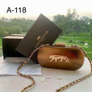 May include: A bronze metallic clutch bag with a gold chain strap and clasp. The bag features a gold panther emblem. The text "A-118" is visible. A black box with "Sabyasachi Calcutta" is in the background. The bag is approximately 20 cm long.