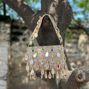 May include: A beige beaded handbag with a rectangular shape and a shoulder strap. The bag is adorned with teardrop-shaped embellishments in various colors, including green, white, and pink. The bag is hanging from a tree branch.