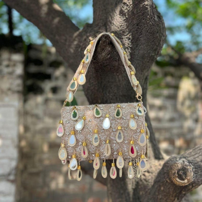 May include: A beige beaded handbag with a rectangular shape and a shoulder strap. The bag is adorned with teardrop-shaped embellishments in various colors, including green, white, and pink. The bag is hanging from a tree branch.