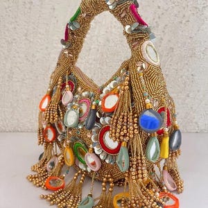 Könnte beinhalten: Eine goldfarbene, mit Perlen besetzte Handtasche mit gebogenem Henkel. Die Tasche ist mit bunten Quasten, Pailletten und dekorativen Spiegeln verziert. Der Griff ist mit grünen, silbernen und goldenen Akzenten versehen. Die Tasche hat eine runde Form und ist mit verschiedenen bunten Perlen verziert.