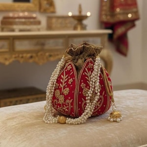 Könnte beinhalten: Ein roter, bestickter Beutel mit goldenen Akzenten und einer Perlenkette. Die Tasche hat florale Muster und eine goldene Quaste. Die Tasche liegt auf einer beigen Oberfläche.