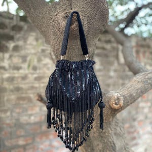 May include: Black beaded handbag with a velvet handle, suspended from a tree branch. The bag has a sequined design and beaded fringe. It features a drawstring closure and two beaded tassels.