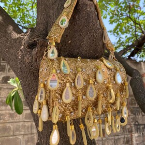 May include: A gold beaded handbag with a shoulder strap. The bag is adorned with teardrop-shaped embellishments and small, clear rhinestones. The bag is hanging from a tree branch.