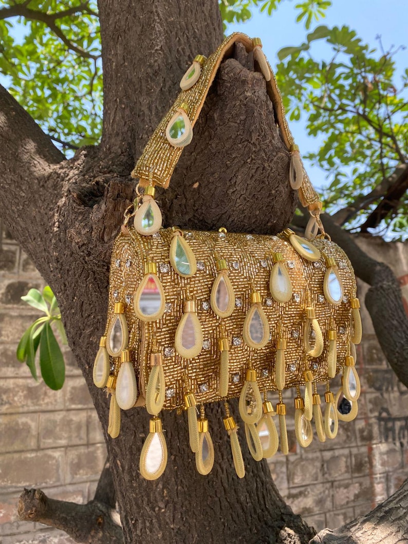 May include: A gold beaded handbag with a shoulder strap. The bag is adorned with teardrop-shaped embellishments and small, clear rhinestones. The bag is hanging from a tree branch.