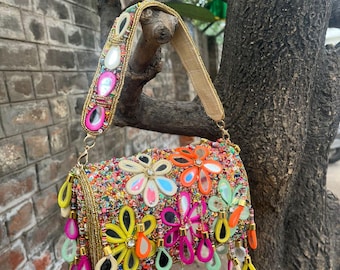 May include: A rectangular handbag featuring a vibrant, multi-colored beaded surface with large, flower-shaped mirror appliqués in yellow, orange, and pink. The bag includes a matching beaded strap and dangling teardrop-shaped mirror charms.