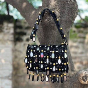 May include: Black beaded handbag with a rectangular shape, adorned with teardrop-shaped embellishments and gold accents. The bag features a black strap and is suspended from a tree branch, showcasing its intricate design and craftsmanship.