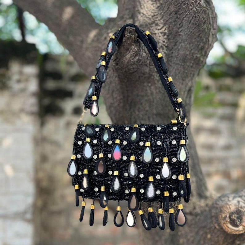 May include: Black beaded handbag with a rectangular shape, adorned with teardrop-shaped embellishments and gold accents. The bag features a black strap and is suspended from a tree branch, showcasing its intricate design and craftsmanship.