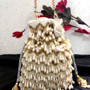 Puede incluir: Un bolso de mano con cuentas de color marfil con una cadena dorada. El bolso está adornado con filas de pequeñas cuentas redondas y cuentas de perlas en forma de lágrima. Los detalles dorados y las borlas añaden detalles a este elegante accesorio.