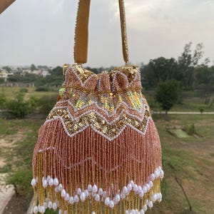Puede incluir: Un bolso de mano con cordón que presenta una sección superior de lentejuelas doradas con un patrón de chevrón, acentuado por cuentas blancas tipo perla. La parte inferior consiste en largos flecos de cuentas color melocotón con cuentas blancas en forma de lágrima.
