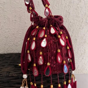 May include: A burgundy velvet drawstring bag with a beaded handle and decorative teardrop-shaped mirrors. The bag features gold accents and dangling mirror and tassel embellishments, creating a glamorous accessory.