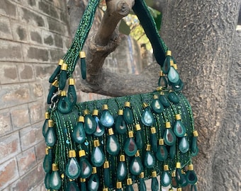 May include: A dark green handbag featuring a dense arrangement of hanging teardrop-shaped charms with reflective centers. The bag is adorned with small green beads and gold-toned metal accents, including a beaded handle and a structured body.