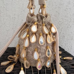 以下が含まれることがあります： ベージュの巾着バッグ。 涙型のミラーアクセントとゴールドカラーの金具が装飾されています。 バッグは柔らかく、質感のある外観で、お揃いのショルダーストラップが付いています。 バッグには、複数の涙型のミラーアクセントが施されています。
