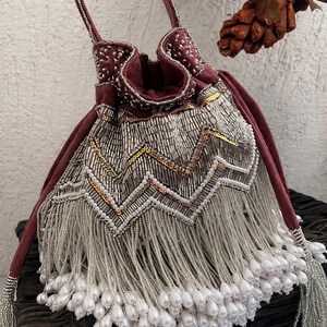 May include: A deep burgundy velvet drawstring pouch bag featuring intricate silver beadwork in a chevron pattern. The lower section is adorned with long silver beaded fringe that terminates in white teardrop-shaped faux pearl beads.