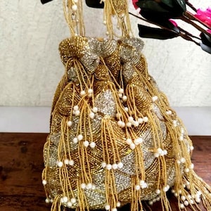 Könnte beinhalten: Eine goldfarbene, perlenbesetzte Handtasche mit Kordelzugverschluss. Die Tasche hat eine Mischung aus goldenen und silbernen Perlen, mit langen Perlenquasten und kleinen weißen Perlenakzenten. Die Griffe sind ebenfalls mit Perlen besetzt.