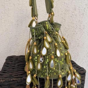 May include: Olive green drawstring bag adorned with sequins and mirror details. The bag has long, teardrop-shaped embellishments with gold-coloured caps and beaded tassels. It features two olive green straps and is set against a neutral backdrop.