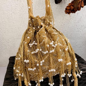 May include: A gold drawstring handbag featuring intricate sequin embroidery in a wavy pattern. The bag is adorned with numerous hanging gold beaded tassels, each tipped with a small white faux pearl. It includes a matching gold beaded handle.