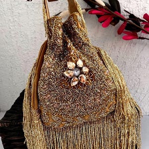 May include: A gold beaded drawstring bag with a suede handle and long fringe. The bag features a floral embellishment with clear and amber-colored crystals. The bag is a warm gold color.