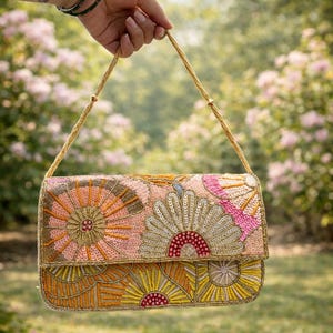 以下が含まれることがあります： ピンク、オレンジ、ゴールドの色合いの花柄が施されたビーズクラッチ。ゴールドのチェーンストラップが付いており、ぼやけた緑と花の背景に持ち上げられています。