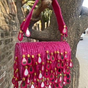 May include: A vibrant hot pink handbag featuring an all-over sequin finish and a single top handle. The bag is adorned with numerous dangling, teardrop-shaped charms that incorporate small mirrors, each secured with a gold-toned metal cap.