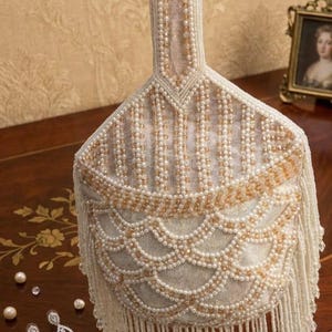 Può includere: Borsa a mano avorio con perline e manico, con un design smerlato e frange. La borsa è adornata con perline bianche e dorate. Un paio di orecchini con perle e diamanti sono sulla superficie di legno.