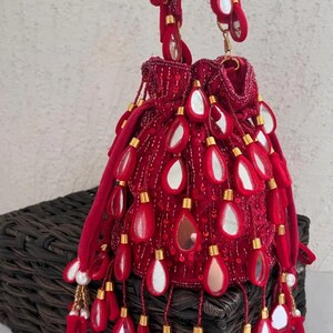 May include: A red velvet drawstring bag adorned with teardrop-shaped mirrors and gold-colored beads. The bag features a beaded handle and decorative tassels, creating an ornate and elegant accessory.