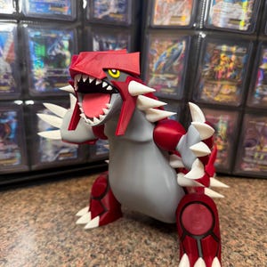 Puede incluir: Una figura impresa en 3D de Groudon, un personaje de Pokémon, en una pose dinámica. La figura presenta una cabeza y acentos rojos, un cuerpo gris y púas blancas. La figura se exhibe frente a una colección de cartas coleccionables.