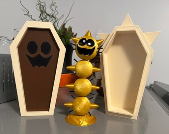 May include: Two white coffin-shaped boxes with a gold and black smiling creature in the middle. One coffin has a black jack-o'-lantern face on the front.