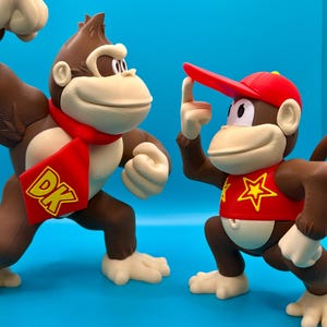 Könnte beinhalten: Zwei braune und hellbraune Donkey Kong Figuren. Eine trägt einen roten Schal mit "DK". Die andere trägt ein rotes Hemd mit einem gelben Stern und eine rote Kappe. Beide haben hellbraune Hände und Füße auf blauem Hintergrund.