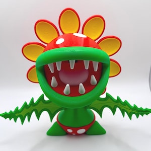 Könnte beinhalten: 3D-gedruckte Figur einer Piranha-Pflanze aus der Super Mario-Videospielserie. Die Pflanze hat einen roten Kopf mit weißen Punkten, einen grünen Mund mit weißen Zähnen, gelbe Blütenblätter und grüne Blätter. Sie trägt rote Shorts mit weißen Punkten.