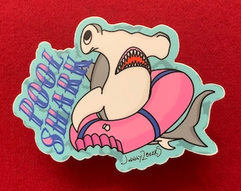 May include: A blue and white sticker with a cartoon hammerhead shark wearing a pink pool float. The sticker says "Pool Shark" in a blue and white font.